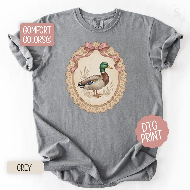 May include: A grey t-shirt with a detailed illustration of a mallard duck in an oval frame with a pink bow and a lace border. The shirt has the words "COMFORT COLORS" and "DTG PRINT" printed on it. The shirt is grey.