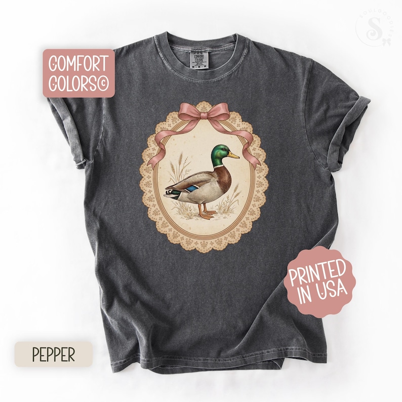 May include: Dark grey Comfort Colors t-shirt with a mallard duck illustration inside an oval frame, adorned with a pink bow. The shirt includes the text "Printed in USA" and "Pepper".