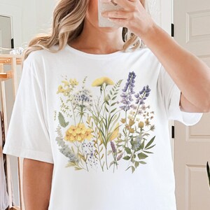 以下が含まれることがあります： 水彩画のフローラルデザインが特徴の白いTシャツ。デザインには、黄色、紫、白のワイルドフラワーと緑の葉が含まれています。シャツは半袖のカジュアルスタイルです。