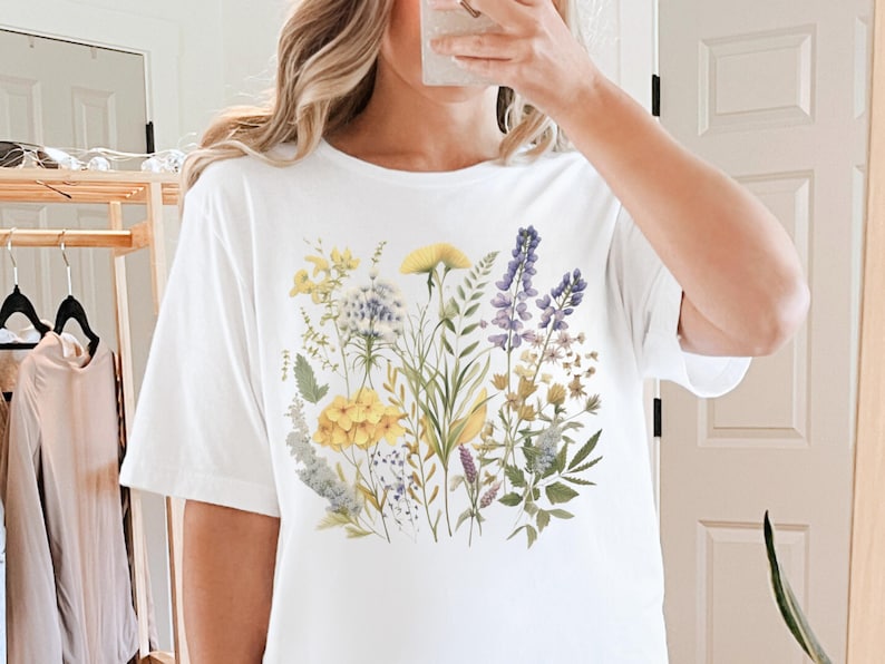以下が含まれることがあります： 水彩画のフローラルデザインが特徴の白いTシャツ。デザインには、黄色、紫、白のワイルドフラワーと緑の葉が含まれています。シャツは半袖のカジュアルスタイルです。