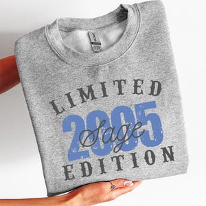 K&ouml;nnte beinhalten: Ein hellgraues Sweatshirt mit dem Text "LIMITED 2005 Sage EDITION" in Blau und Dunkelgrau. Das Sweatshirt wird von einer Person gehalten. Das Produkt ist mit "SWEATER - SP.GREY" gekennzeichnet.