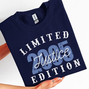 K&ouml;nnte beinhalten: Marineblaues Sweatshirt mit dem Text "LIMITED 2005 Justice EDITION" in Wei&szlig; und Blau. Das Sweatshirt wird von einer Person gehalten. Die Worte "SWEATER - NAVY" sind vertikal auf der rechten Seite aufgedruckt.