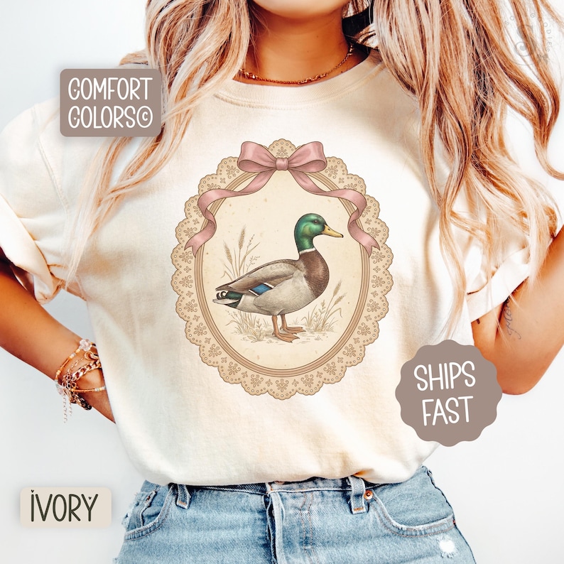 Könnte beinhalten: Elfenbeinfarbenes T-Shirt mit einer detaillierten Illustration einer Stockente in einem ovalen Rahmen, verziert mit einer rosa Schleife. Der Rahmen hat ein spitzenartiges Design. Der Text "COMFORT COLORS" und "SHIPS FAST" sind ebenfalls sichtbar.