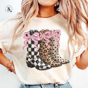 Könnte beinhalten: Cremefarbenes T-Shirt mit drei Cowboy-Stiefeln mit rosa Schleifen. Die Stiefel haben verschiedene Muster: schwarz-weißes Schachbrettmuster, Leopardenmuster und ein beiges Polka-Dot-Design. Eine lustige, stilvolle Grafik.