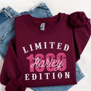 K&ouml;nnte beinhalten: Burgunderfarbenes Sweatshirt mit dem Text "LIMITED 1999 Harley EDITION" in wei&szlig;er und rosa Schrift. Das Sweatshirt liegt gefaltet auf einer blauen Jeans.