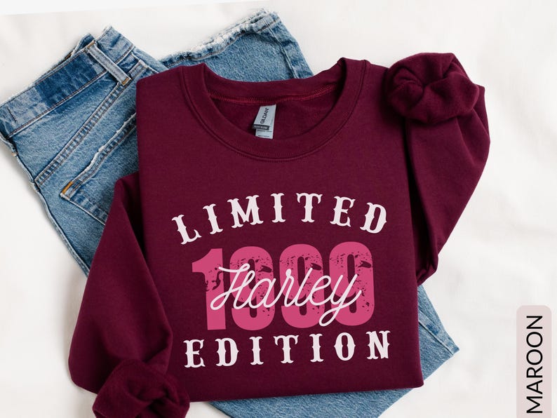 K&ouml;nnte beinhalten: Burgunderfarbenes Sweatshirt mit dem Text "LIMITED 1999 Harley EDITION" in wei&szlig;er und rosa Schrift. Das Sweatshirt liegt gefaltet auf einer blauen Jeans.