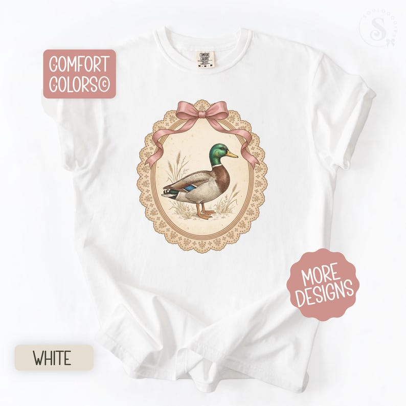 May include: White t-shirt with a vintage-style illustration of a mallard duck in an oval frame. The frame has a pink bow and a lace-like border. The words "Comfort Colors" and "More Designs" are also visible.
