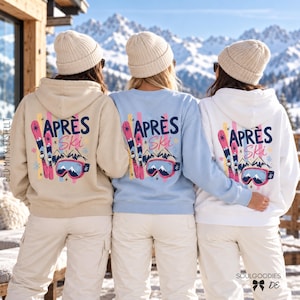Op de afbeelding: Drie personen in beige, lichtblauwe en witte hoodies en gebreide mutsen. De achterkant van de kledingstukken is voorzien van een afbeelding met de tekst APRÈS Ski, roze ski's, een skibril en illustraties van bergen.