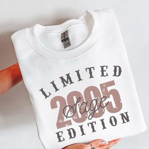 K&ouml;nnte beinhalten: Wei&szlig;es Sweatshirt mit dem Text "LIMITED 2005 Sage EDITION" in braunen und grauen Buchstaben. Das Sweatshirt wird von einer Person gehalten. Die Worte "SWEATER WHITE" sind vertikal auf der rechten Seite aufgedruckt.