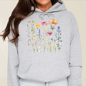 May include: A light grey hooded sweatshirt featuring a watercolour-style graphic print of various wildflowers in shades of yellow, pink, orange, and blue. The garment includes a front kangaroo pocket and a matching grey drawstring at the neckline.