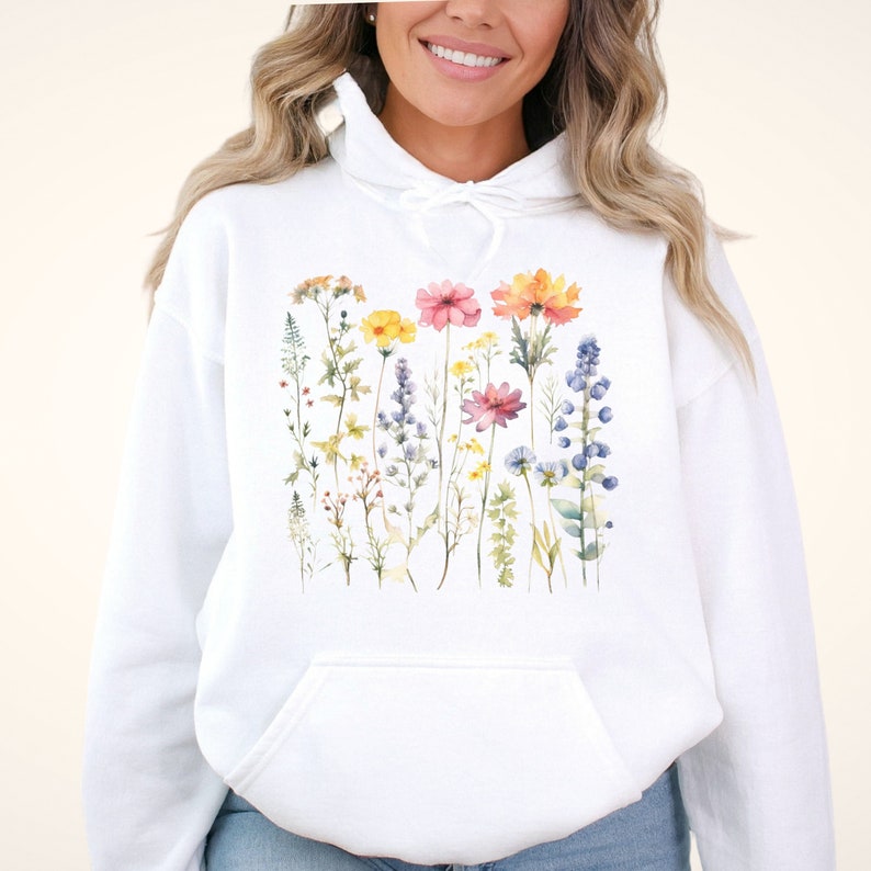 May include: A white hooded sweatshirt featuring a central graphic print of various wildflowers in watercolour style. The design includes yellow, pink, blue, and orange floral stems and leaves arranged in a vertical pattern across the chest area.