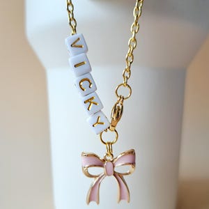 Könnte beinhalten: Eine goldene Kettenkette mit einem rosa Schleifenanhänger und weißen Buchstabenperlen, die "VICKY" buchstabieren.