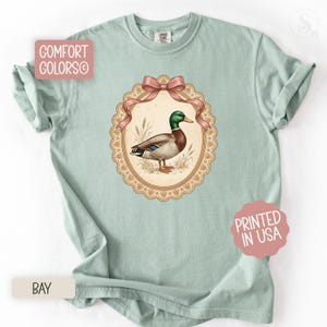 May include: A pale green t-shirt with a framed illustration of a mallard duck. The duck is brown, green, and blue, set against a beige background with a pink bow. The shirt has the text "COMFORT COLORS" and "PRINTED IN USA".