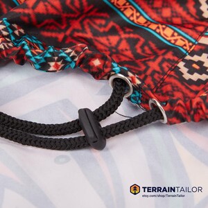 May include: Close-up of a patterned fabric with a drawstring. The fabric features a vibrant design in red, black, turquoise, and orange. A black drawstring with a plastic cord lock is visible, along with metal grommets.