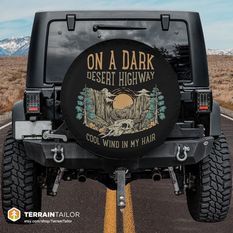 May include: Black spare tire cover with a desert landscape design. The cover features text that reads "ON A DARK DESERT HIGHWAY COOL WIND IN MY HAIR". The design includes a skull, sun, and trees.