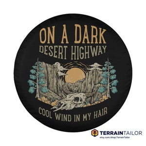 May include: Black spare tire cover with a desert landscape design. The cover features a mountain scene with a setting sun, a skull, and text that reads "ON A DARK DESERT HIGHWAY COOL WIND IN MY HAIR". The Terrain Tailor logo is at the bottom.