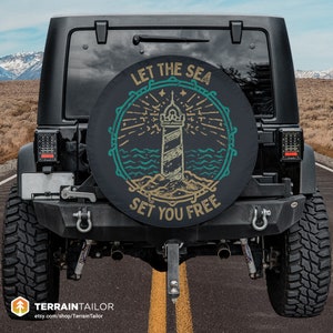 May include: A black Jeep with a spare tire cover that says "Let the Sea Set You Free" in a vintage style font. The cover features a lighthouse with a blue and green ocean scene.