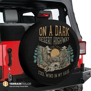 May include: Black spare tire cover with a desert landscape design. The cover features the text "ON A DARK DESERT HIGHWAY COOL WIND IN MY HAIR" with a skull, sun, and mountain graphic. The cover is mounted on a red Jeep.