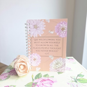 May include: A spiral-bound notebook with a floral design and the quote "LIKE WILDFLOWERS; YOU MUST ALLOW YOURSELF TO GROW IN ALL THE PLACES PEOPLE THOUGHT YOU NEVER WOULD." The notebook is peach-colored with white and purple flowers.