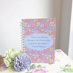 May include: A spiral-bound notebook with a floral pattern in purple, orange, and green. The cover features a quote by Henri Matisse: "There are always flowers, for those who want to see them." Decorative flowers in green and lavender are next to the notebook.