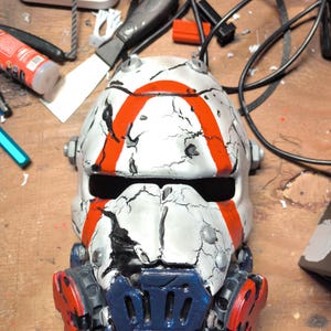 May include: A white helmet with a distressed, cracked finish and bold orange stripes. The helmet features a dark blue, vented faceplate section at the bottom, integrated side components with red accents, and a horizontal eye opening across the front.