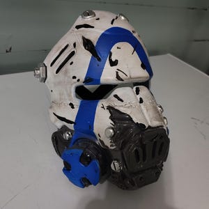 May include: A white and blue helmet with black accents. The helmet has a blue stripe and a black lower section. Silver rivets are visible on the helmet's surface. The helmet appears to be a prop or costume piece.