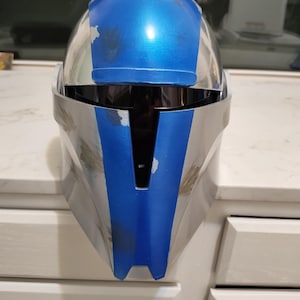 May include: A silver and blue helmet with a visor. The helmet has a blue top and a silver base. The helmet is sitting on a white surface.