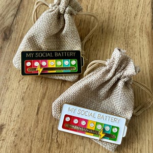 May include: Two rectangular enamel pins labelled MY SOCIAL BATTERY, each featuring a colourful battery level indicator with emoji faces ranging from sad to happy and a yellow lightning bolt slider, resting on small beige drawstring burlap pouches.