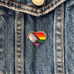 May include: A heart-shaped enamel pin with a rainbow pride flag design. The pin says "You can be yourself with pride" around the edge.