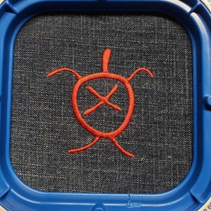 May include: A denim fabric is stretched within a blue embroidery hoop. A red embroidered turtle design is centered on the fabric. The hoop has a warning label and the text "Strong Magnets".