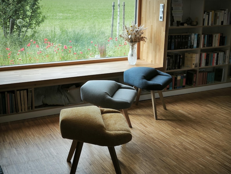 May include: Three modern accent chairs with wooden legs and curved, upholstered seats in mustard yellow, gray, and blue. The chairs are arranged in a room with a large window overlooking a green field.