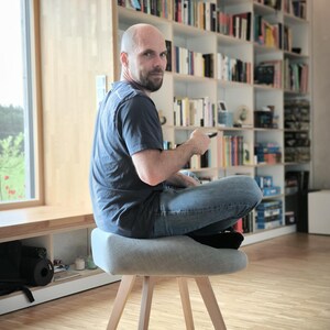 May include: A light gray upholstered stool with wooden legs. The stool has a rounded seat and a simple, modern design. The legs are angled outwards for stability. The stool is in a room with a bookshelf and a window.