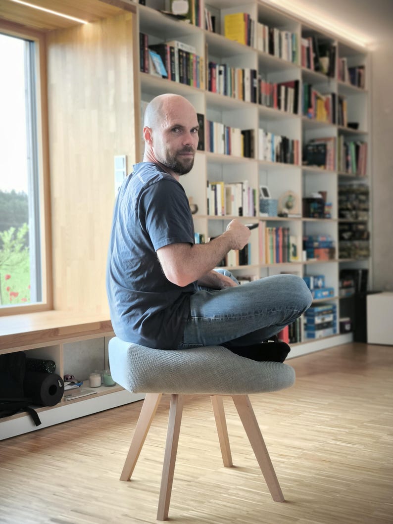 May include: A light gray upholstered stool with wooden legs. The stool has a rounded seat and a simple, modern design. The legs are angled outwards for stability. The stool is in a room with a bookshelf and a window.