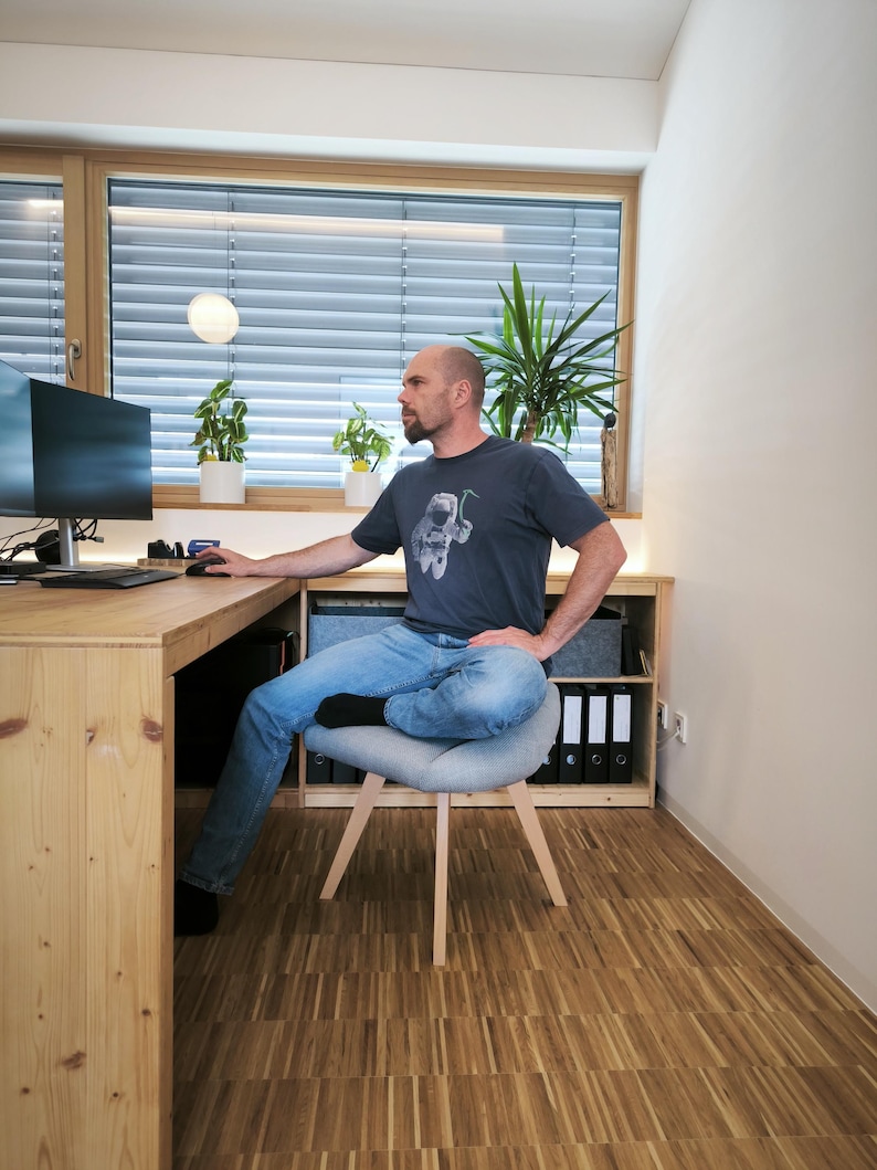 May include: A man sits at a wooden desk in a home office. He wears a dark gray t-shirt and blue jeans. A gray chair with wooden legs is positioned in front of a wooden cabinet. The room has a large window with blinds and potted plants.
