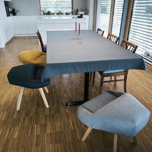 May include: A dining room scene featuring a large rectangular table covered with a gray tablecloth. Several chairs of different designs surround the table. The room has wooden floors and large windows with blinds.