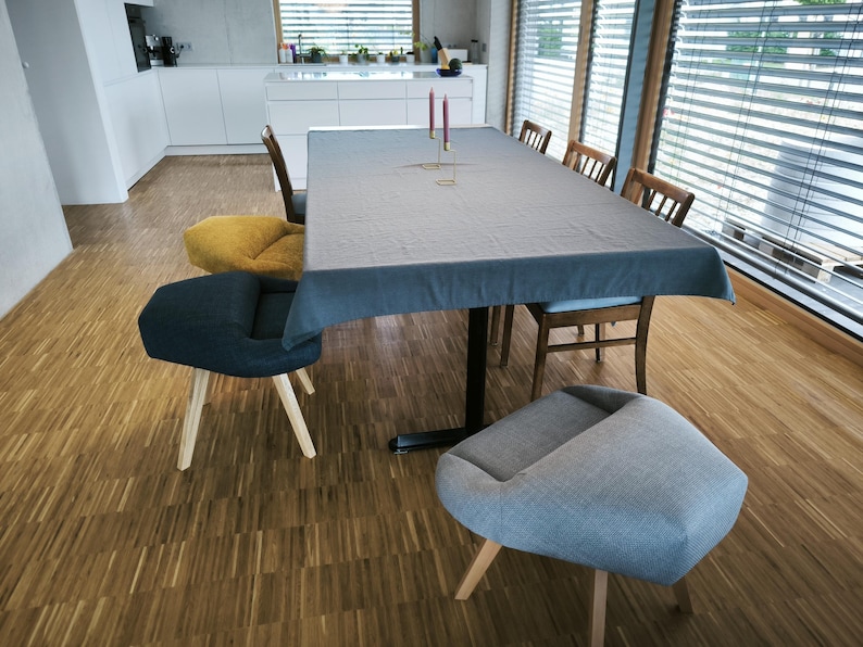 May include: A dining room scene featuring a large rectangular table covered with a gray tablecloth. Several chairs of different designs surround the table. The room has wooden floors and large windows with blinds.
