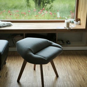 May include: A modern, gray upholstered chair with a rounded seat and three wooden legs. The chair is positioned on a hardwood floor, with a window and a bookshelf in the background. The chair's design is minimalist and contemporary.