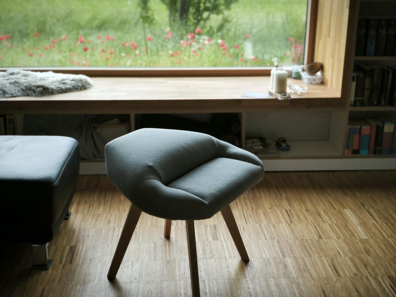 May include: A modern, gray upholstered chair with a rounded seat and three wooden legs. The chair is positioned on a hardwood floor, with a window and a bookshelf in the background. The chair's design is minimalist and contemporary.