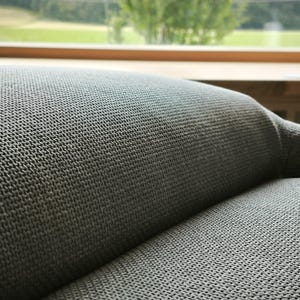 May include: Close-up of a section of a gray upholstered sofa. The fabric has a textured, woven appearance. The background shows a window with a view of green trees and a wooden sill.