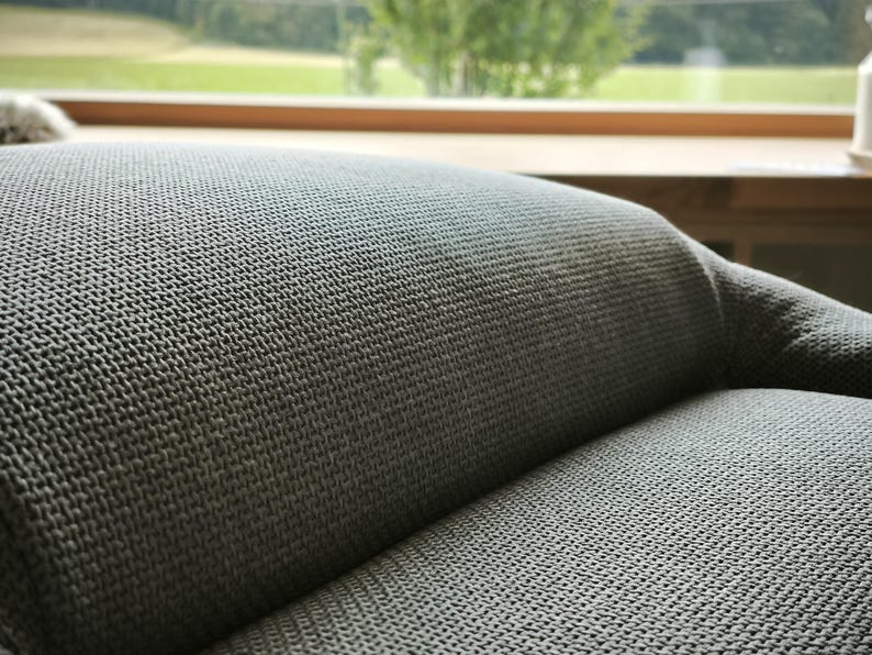 May include: Close-up of a section of a gray upholstered sofa. The fabric has a textured, woven appearance. The background shows a window with a view of green trees and a wooden sill.