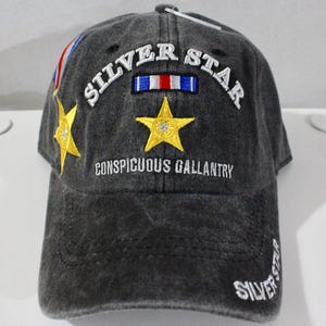 May include: A dark gray baseball cap with the words "SILVER STAR" embroidered in white. The cap features a gold star with a rhinestone center and the text "CONSPICUOUS GALLANTRY". A small tag is attached to the cap.