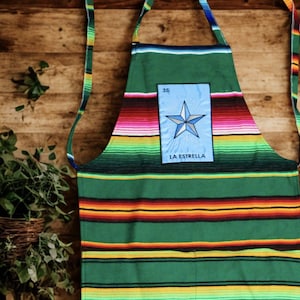 May include: Green apron with colourful horizontal stripes and a blue patch featuring a star and the text "LA ESTRELLA". The apron has a neck strap and waist ties, ideal for cooking or crafting.