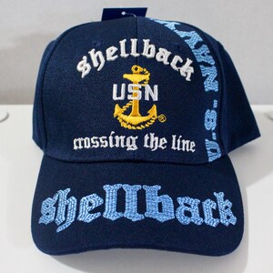 Peut inclure: Casquette de baseball bleu marine avec texte brodé et un emblème d'ancre doré. La casquette présente les mots "Shellback", "USN" et "crossing the line". Le bord a également le mot "Shellback" brodé en bleu.