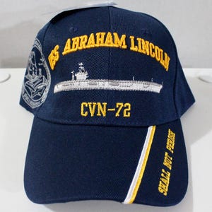 Puede incluir: Gorra de béisbol azul marino con texto bordado en oro "USS Abraham Lincoln" y "CVN-72". La gorra presenta una franja blanca y amarilla en la visera y las palabras "Shall Not Perish".