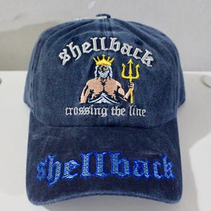 Peut inclure: Casquette de baseball bleu marine avec le mot "Shellback" brodé en blanc au-dessus d'un dessin d'un homme couronné avec un trident. L'expression "crossing the line" est en dessous. Le mot "Shellback" est brodé sur la visière.