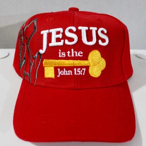 May include: A red baseball cap with the words "JESUS is the" and "John 15:7" embroidered in white and yellow. The cap features a key design and a decorative pattern on the side.