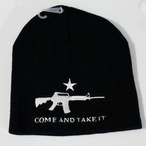 May include: A black knit beanie featuring white embroidery of a rifle and a single star above it. Below the rifle, the text "COME AND TAKE IT" is embroidered in white. A small clear plastic hanging hook is attached to the top of the beanie.