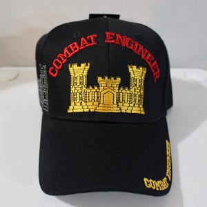 Op de afbeelding: Zwarte baseballcap met "COMBAT ENGINEER" geborduurd in rood en goud. De cap heeft een gouden kasteelembleem en extra tekst op de zijkant en rand. Een klassieke, verstelbare pet.