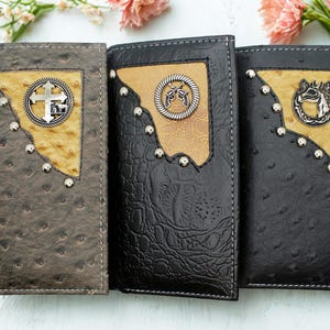 May include: Three leather wallets with decorative metal accents. The wallets are in shades of brown and black, with a textured finish. Each wallet features a different metal emblem and stud details.