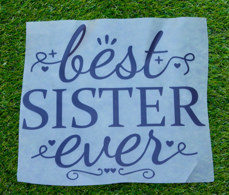 May include: A translucent sheet with the words "best SISTER ever" in a dark blue, cursive font. The text is surrounded by decorative elements like hearts and flourishes, set against a backdrop of green grass.
